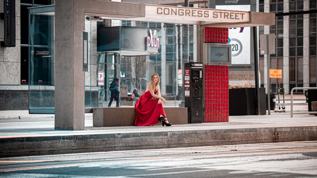 A stylish woman in a red dress sits at a Detroit bus stop on Congress Street.