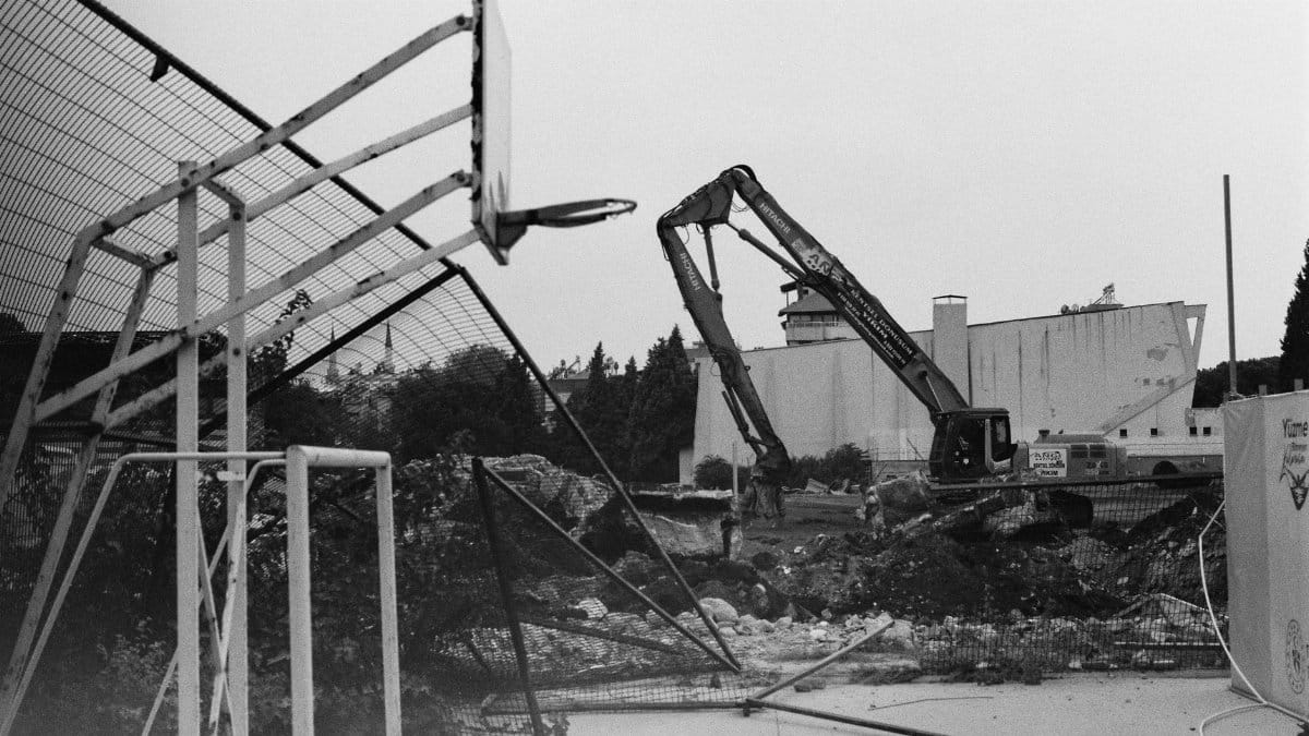 Black and white image of an urban demolition site featuring a dismantled basketball hoop and heavy machinery.