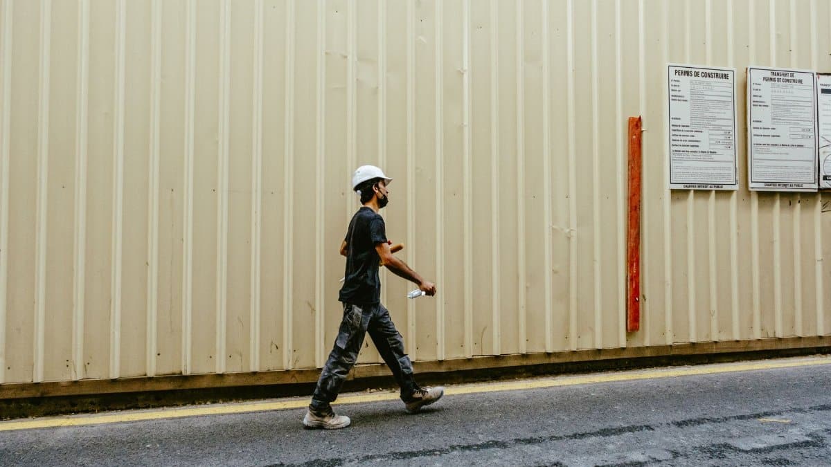 An adult male construction worker walks along an outdoor construction site wall.