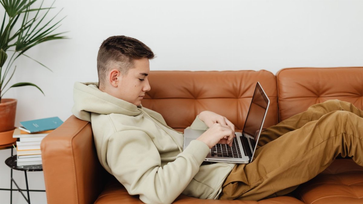 Teenager lounging on a couch while using a laptop, surrounded by books at home.