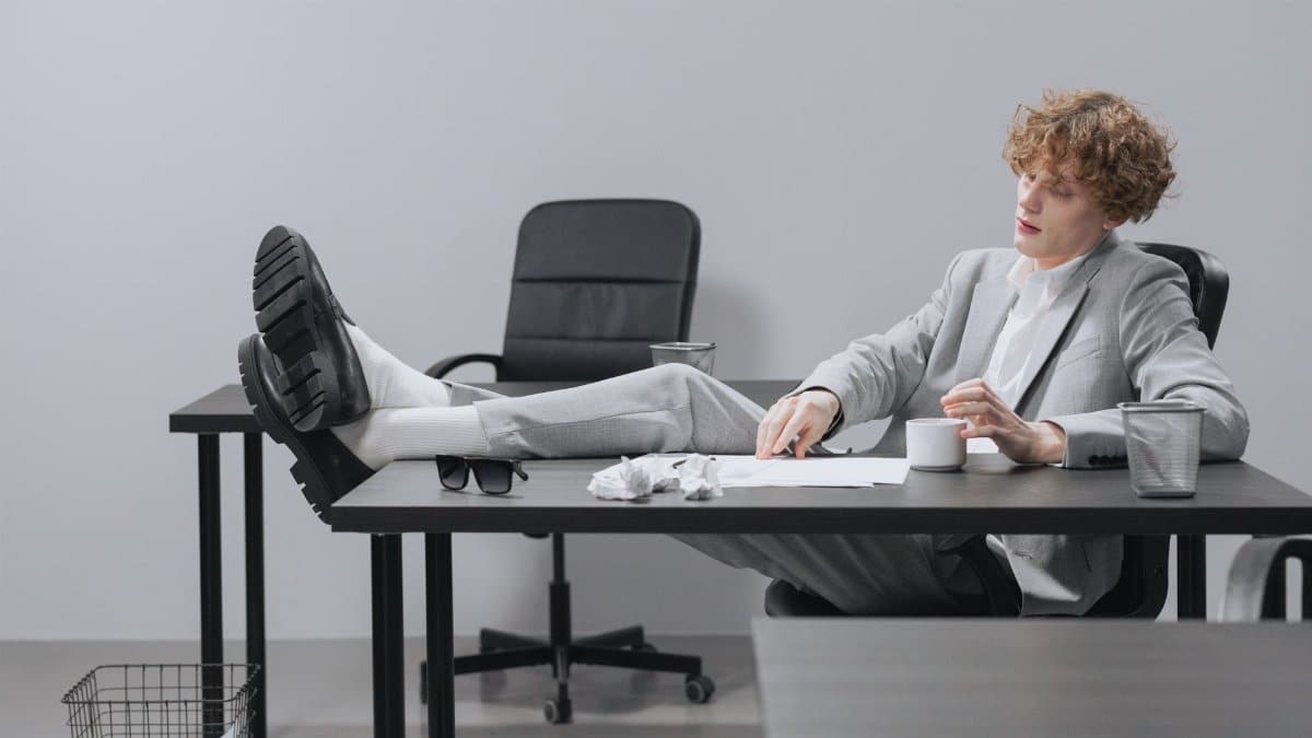 A curly-haired man in a suit relaxes with feet on desk, looking serious in a modern office setting.