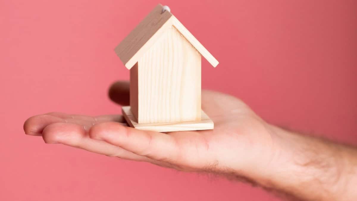 Wooden house model in hand symbolizing real estate concepts on pink background.