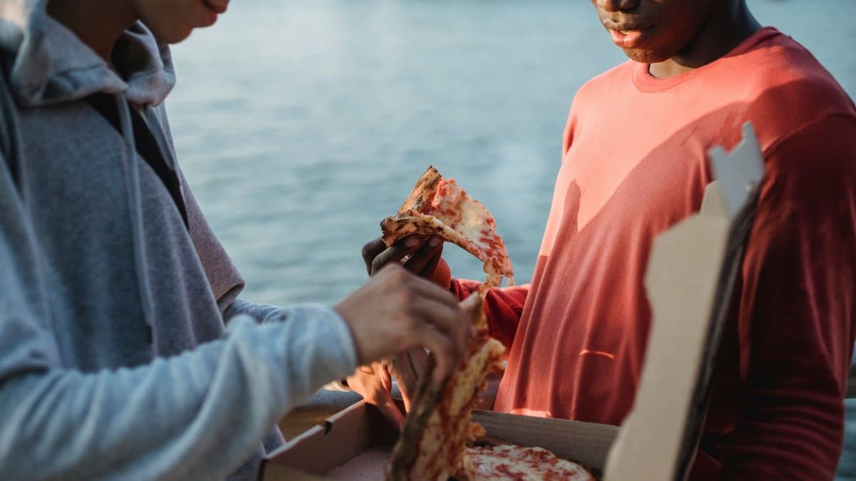 Crop diverse male friends standing on city embankment and enjoying fresh appetizing pizza in evening sun