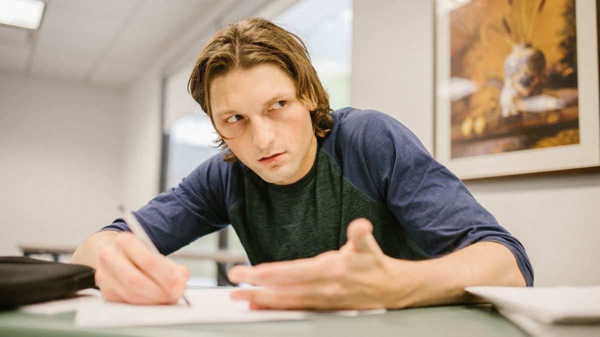 Male college student looking away with a cheat sheet during an exam in a classroom setting.