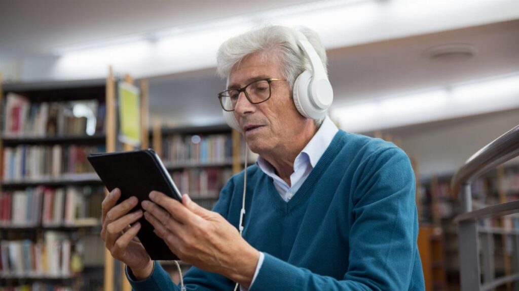 Senior man in a library using a tablet and headphones, enjoying digital media.