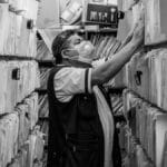 Elderly man sorts through files in an office aisle wearing a face mask.