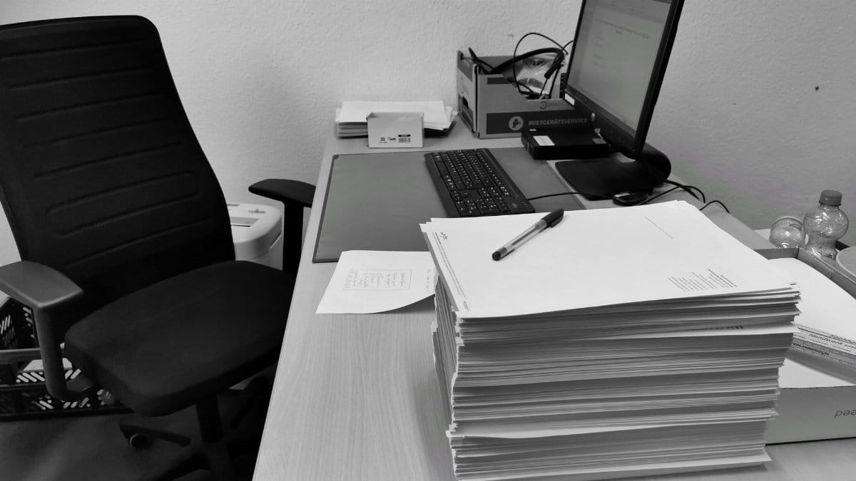 Black and white image of a cluttered office desk with a computer, chair, and paperwork.