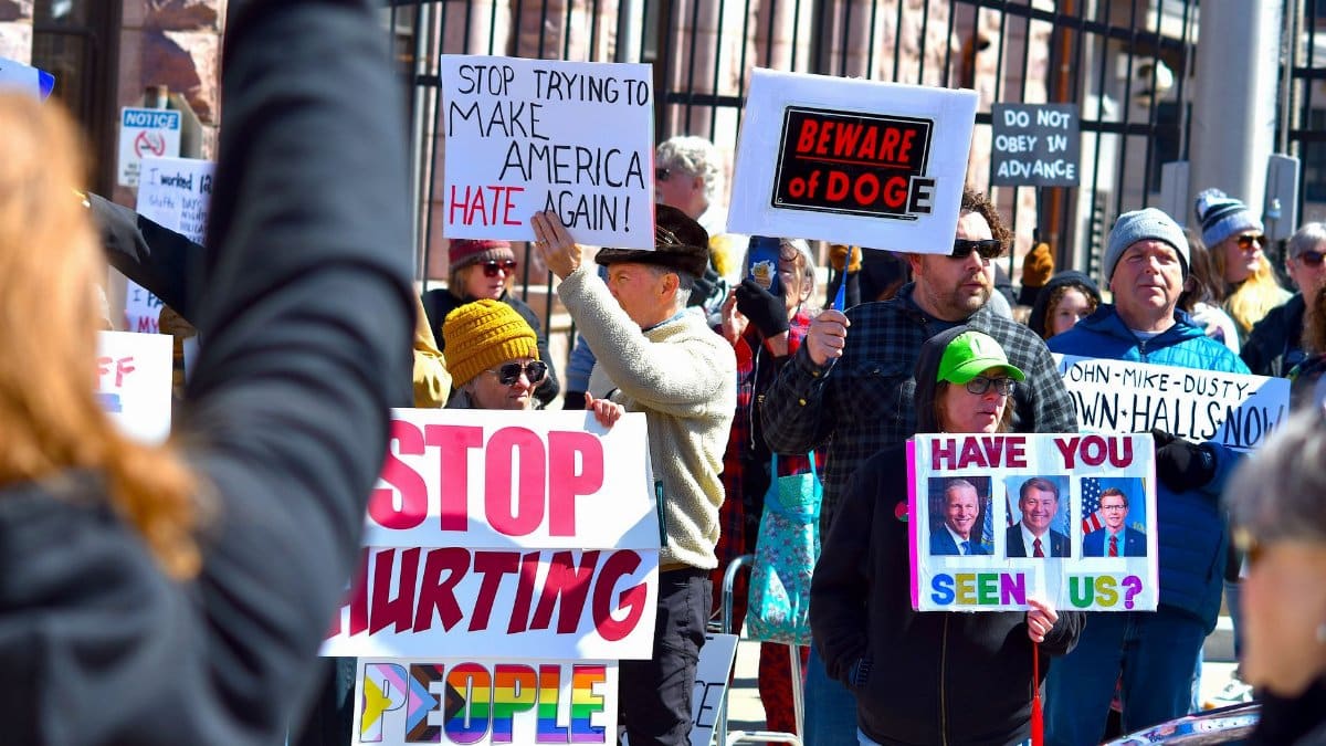 Crowd at a protest holding various signs with political messages in daylight.