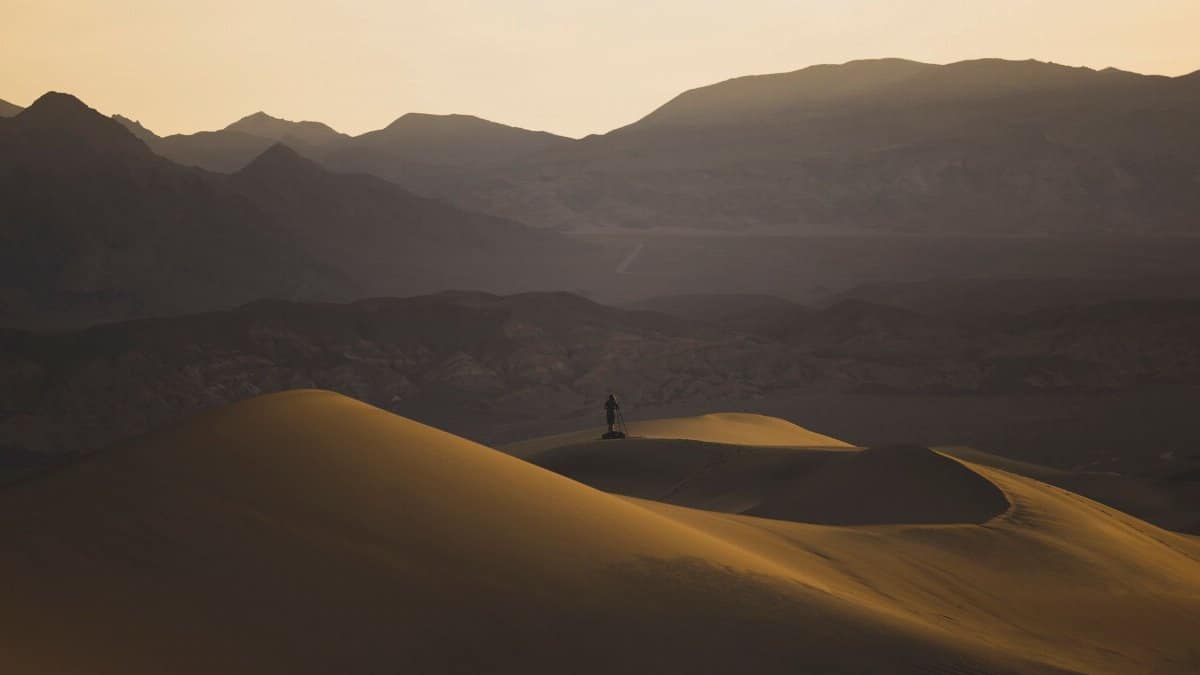 A lone figure stands atop sunlit sand dunes with mountains in the background at sunset.