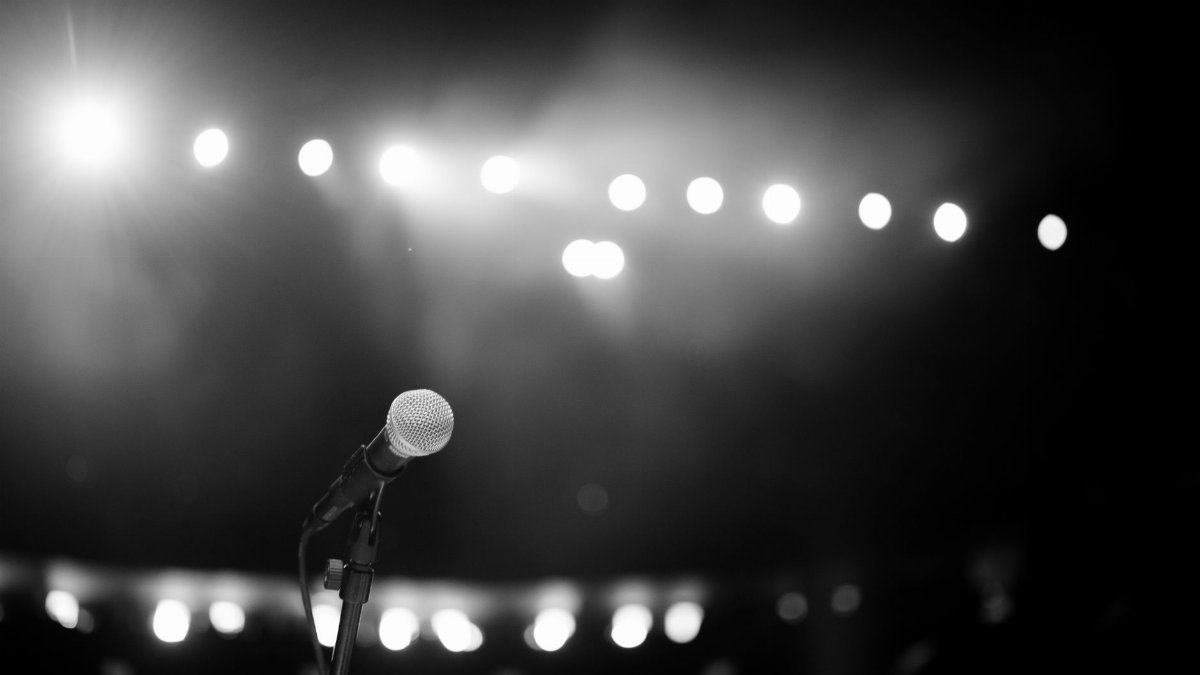 A dramatic black and white photo of a microphone onstage with bright concert lights.