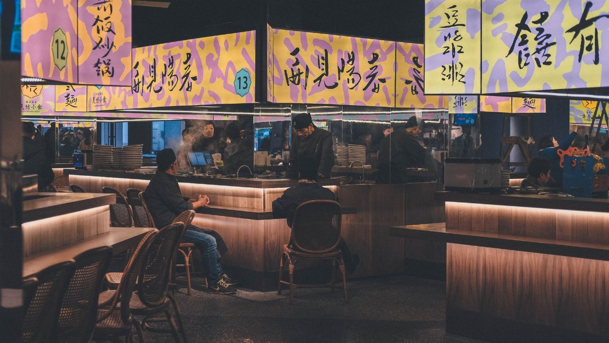 Nighttime view of a bustling Asian food market with colorful signage and diners.