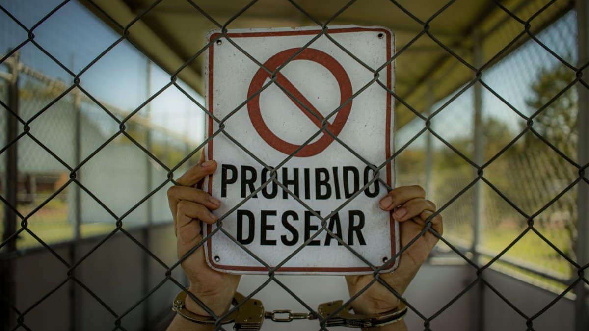 Handcuffed hands hold a 'Prohibido Desear' sign behind a chain-link fence, emphasizing restriction.