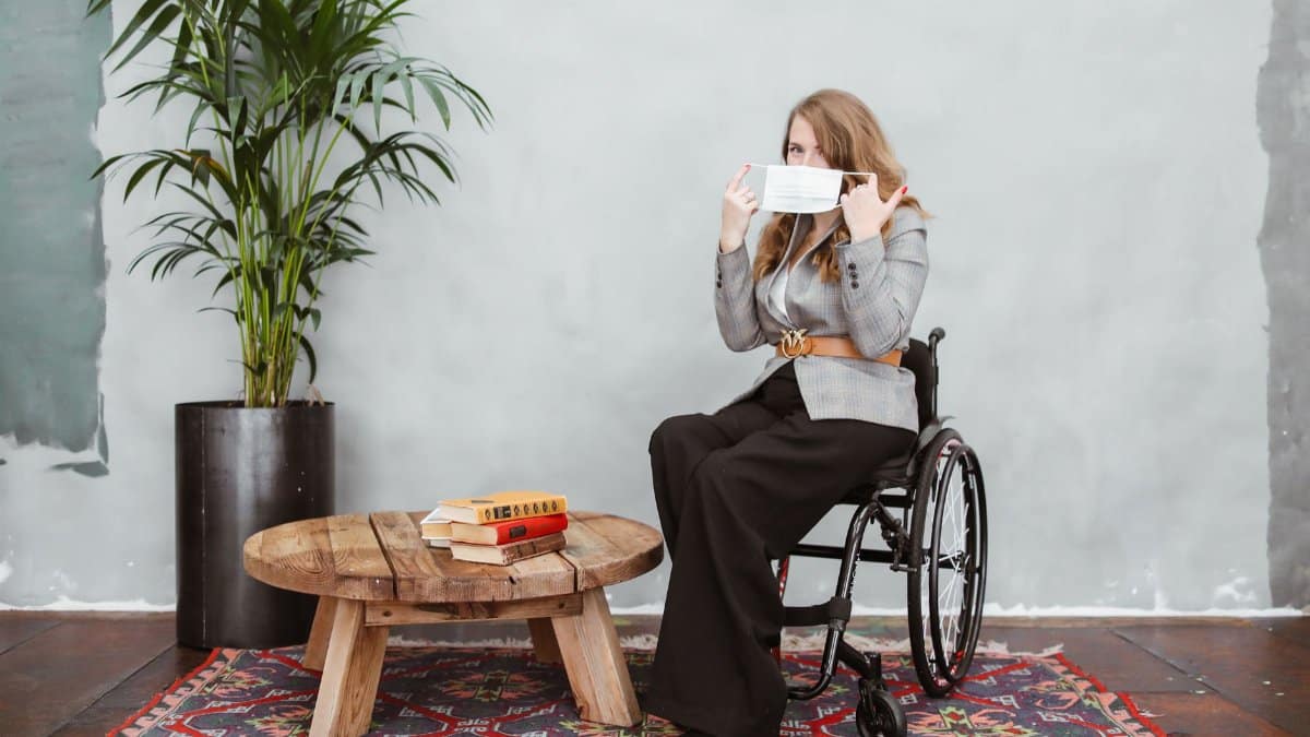 A woman in a wheelchair adjusts her face mask while sitting indoors, emphasizing safety and disability awareness.