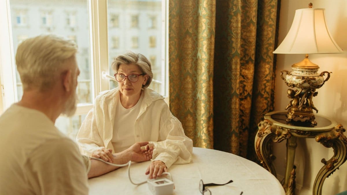 Senior woman checks man's blood pressure at home. Warm light, caring interaction.
