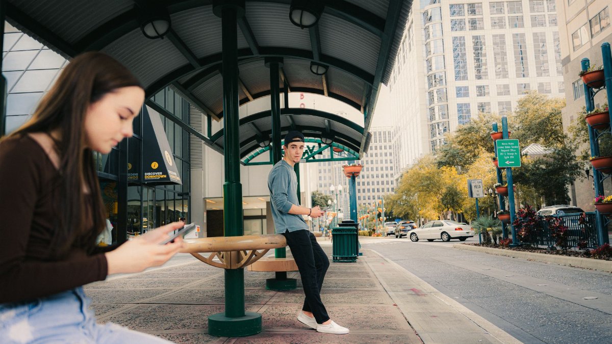 Teens using mobile phones at a city bus stop, urban setting with modern architecture.