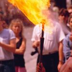 Shirtless street performer breathing fire in a lively Paris scene with a captivated audience.