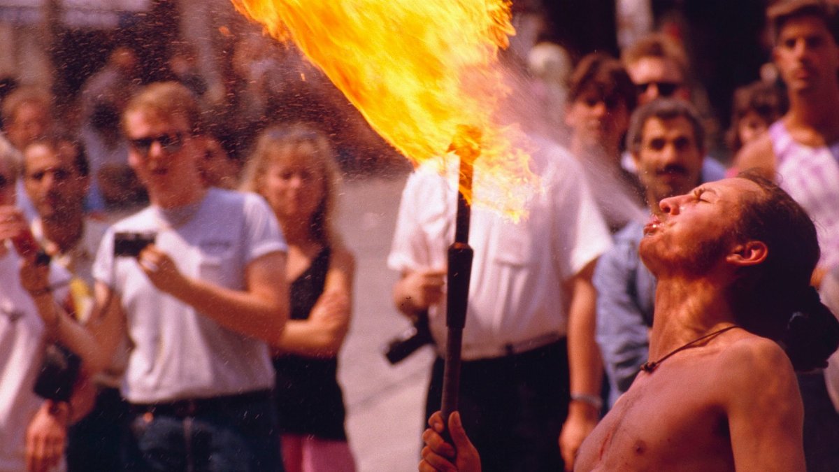 Shirtless street performer breathing fire in a lively Paris scene with a captivated audience.