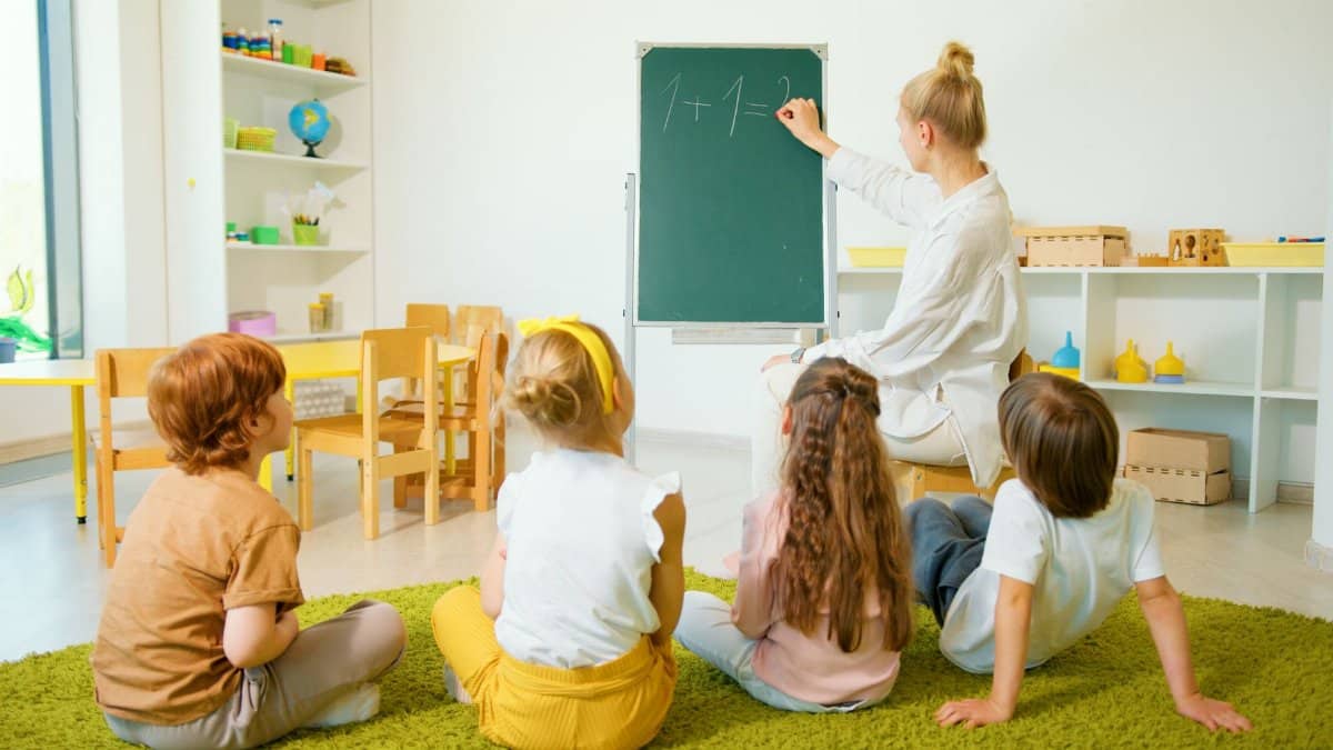 Teacher instructing young students with math on a chalkboard in a bright classroom setting.