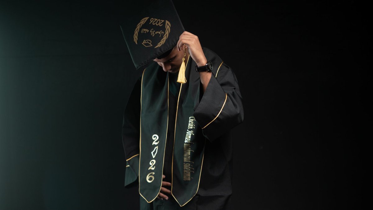 A graduate wearing a cap and gown, class of 2026, looking down in a studio setting.