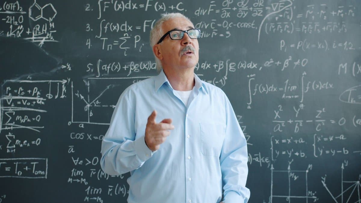 Senior male professor explaining mathematical formulas on a chalkboard.