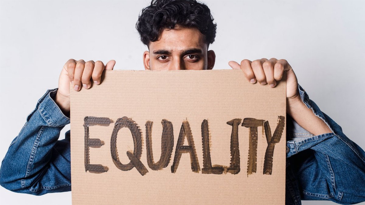 A man holds a cardboard sign reading 'EQUALITY' in a studio.