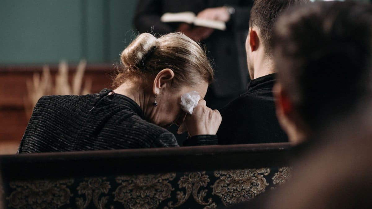 A grieving woman wipes tears during a heartfelt funeral ceremony in a somber indoor setting.