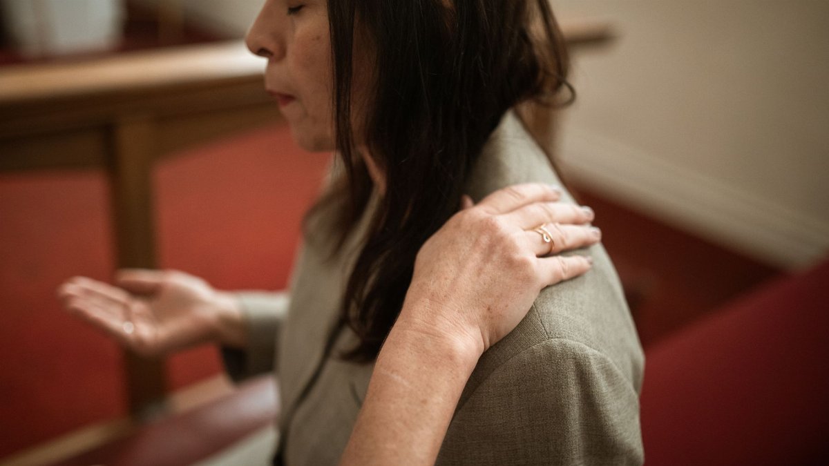 A woman prays indoors, with another person's hand on her shoulder for comfort, expressing faith and spirituality.
