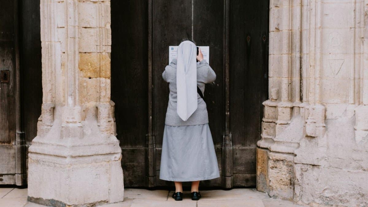 A nun reads a notice on a large wooden church door, surrounded by historic stone architecture.