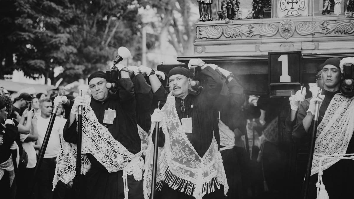 Men in traditional attire carry an altar during a religious procession with a crowd in the background.