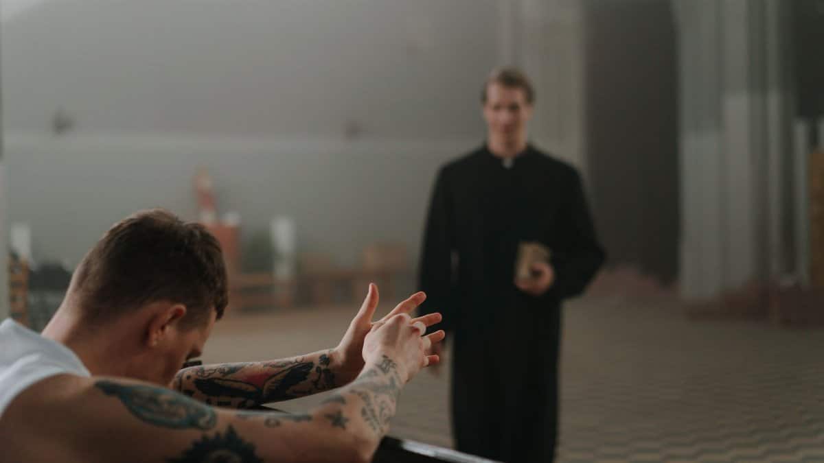 A tattooed man prays in a church as a priest observes, capturing faith and reflection indoors.