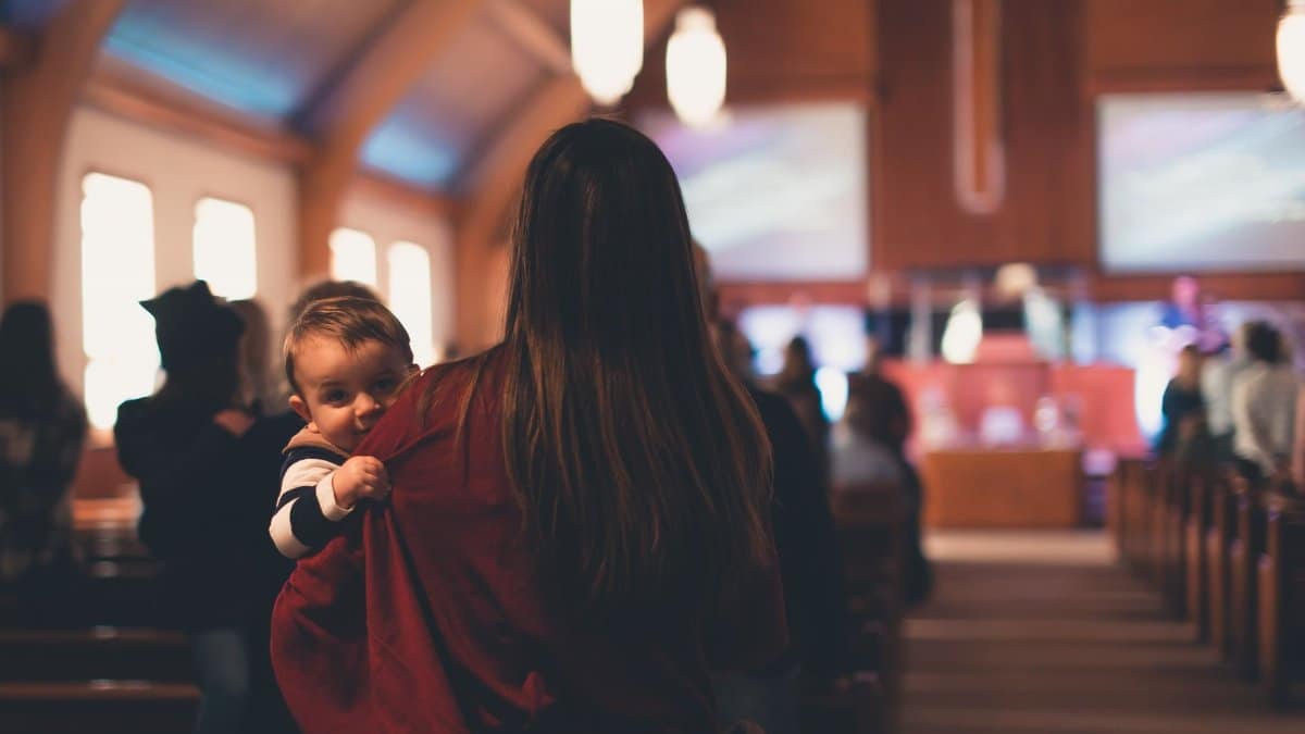 Woman carrying a child inside a church with people worshipping in the background.