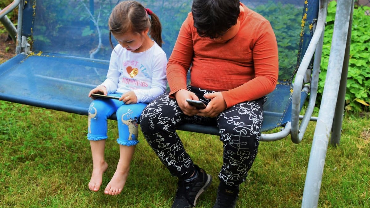 Two children using mobile phones while sitting on a swing outdoors in a garden.