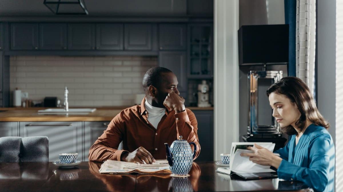 A couple sits at a dining table in a modern kitchen, reading and relaxing.