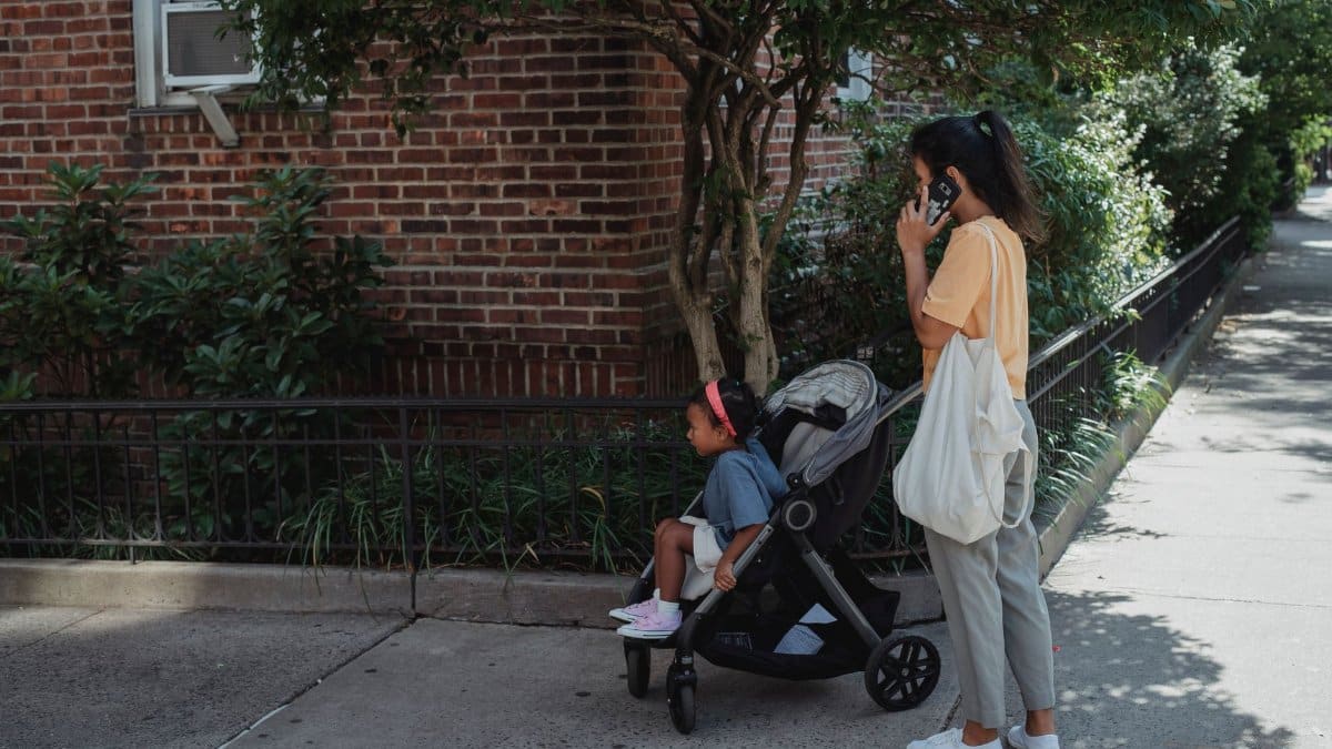 Full body side view of ethnic mother with bag having phone call while standing on sidewalk with girl in carriage near brick building on street