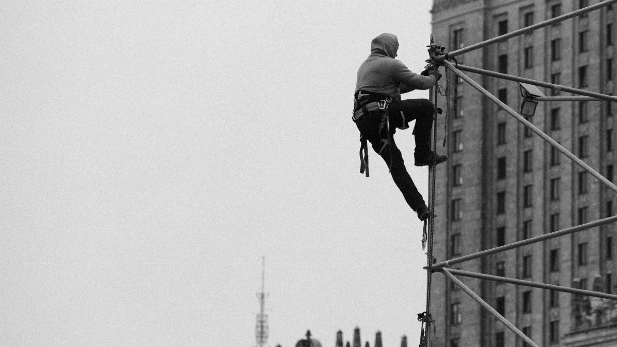A person in safety gear climbs scaffolding in an urban setting, showcasing construction work.