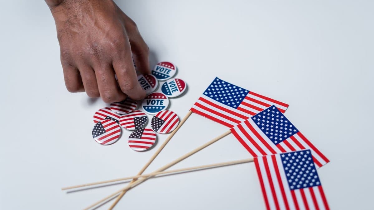 A hand reaches for voting buttons and American flags on a white background.