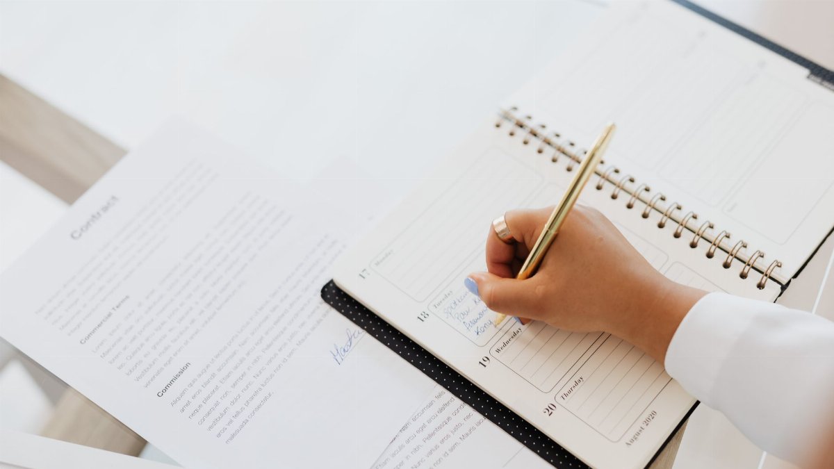 Close-up of a person writing in a planner next to a contract document on a desk.