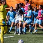 A women's soccer team celebrating after scoring a goal during a daytime match.