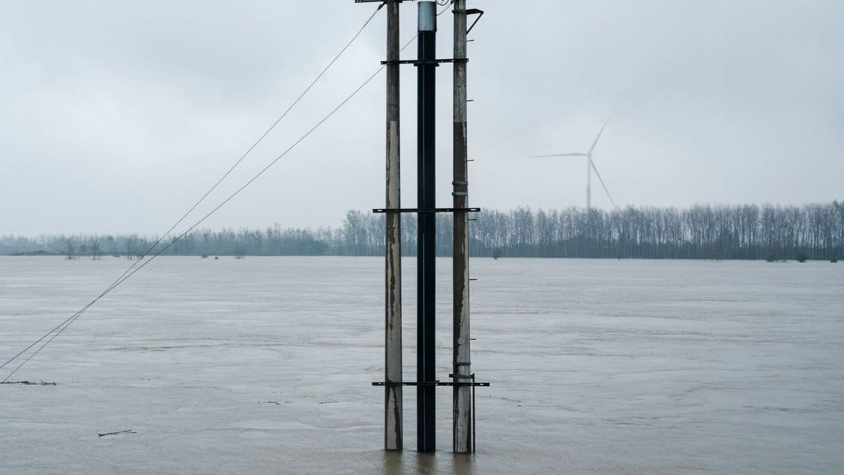 Flooded river landscape featuring a solar panel tower and distant wind turbine under overcast skies.