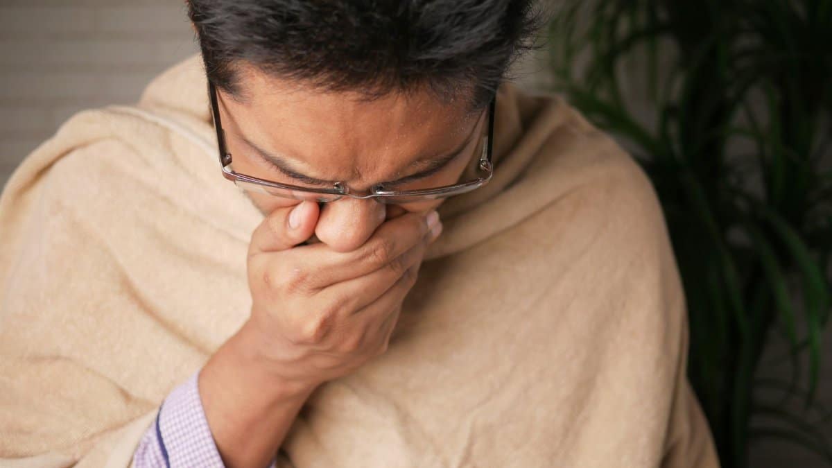 Adult wearing glasses coughs into hand indoors, indicating illness or flu symptoms.