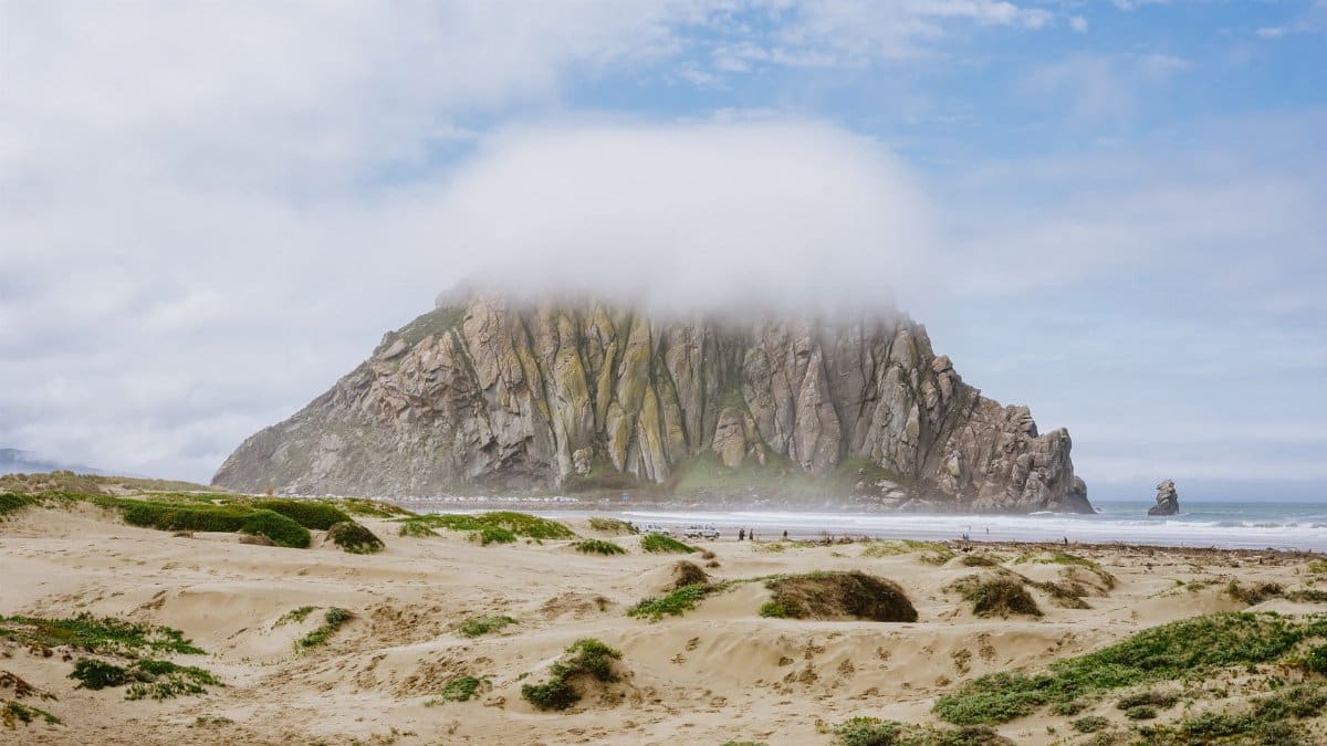 Morro Rock shrouded in fog with sandy beach and sea in Morro Bay, CA.