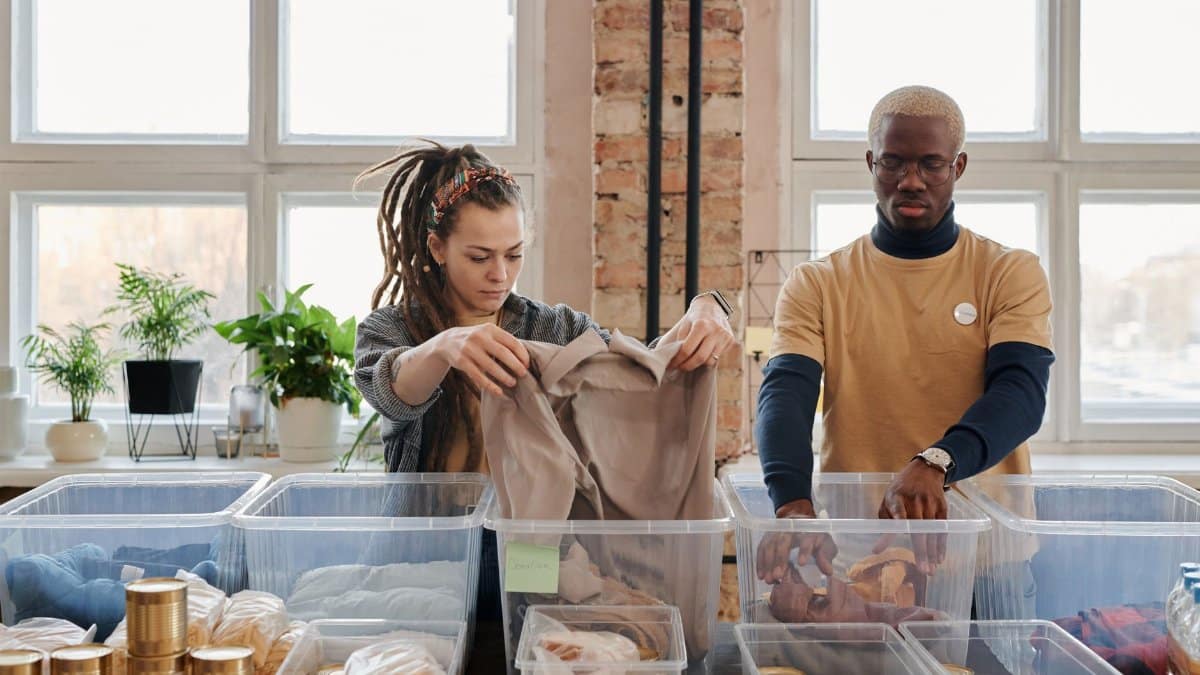 Two volunteers organize clothing and food donations indoors during daylight.