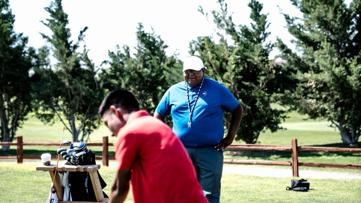 Two golfers on a sunny golf course, one practicing and a caddie observing.