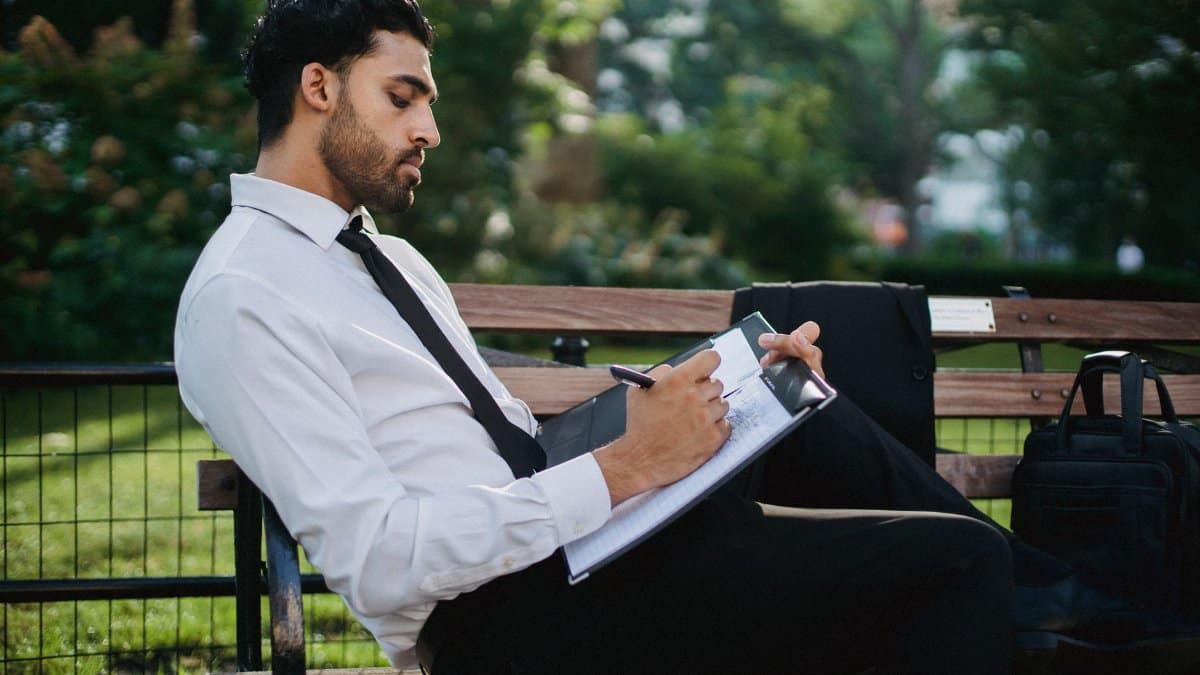 A thoughtful businessman writing notes on a clipboard while sitting on a park bench.