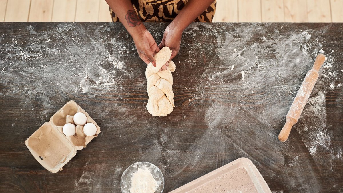 Hands kneading braided dough on a floured wooden table. Includes eggs and a rolling pin.
