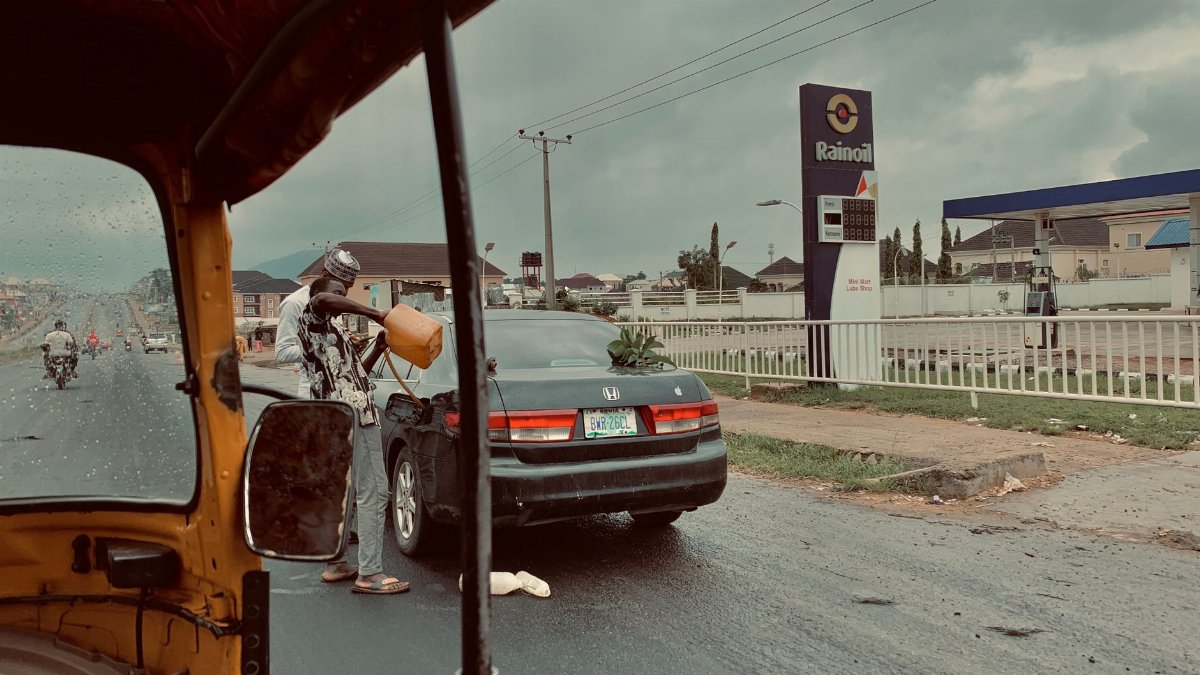 A man fills petrol from a container into a car by a Rainoil station, on a rainy street.