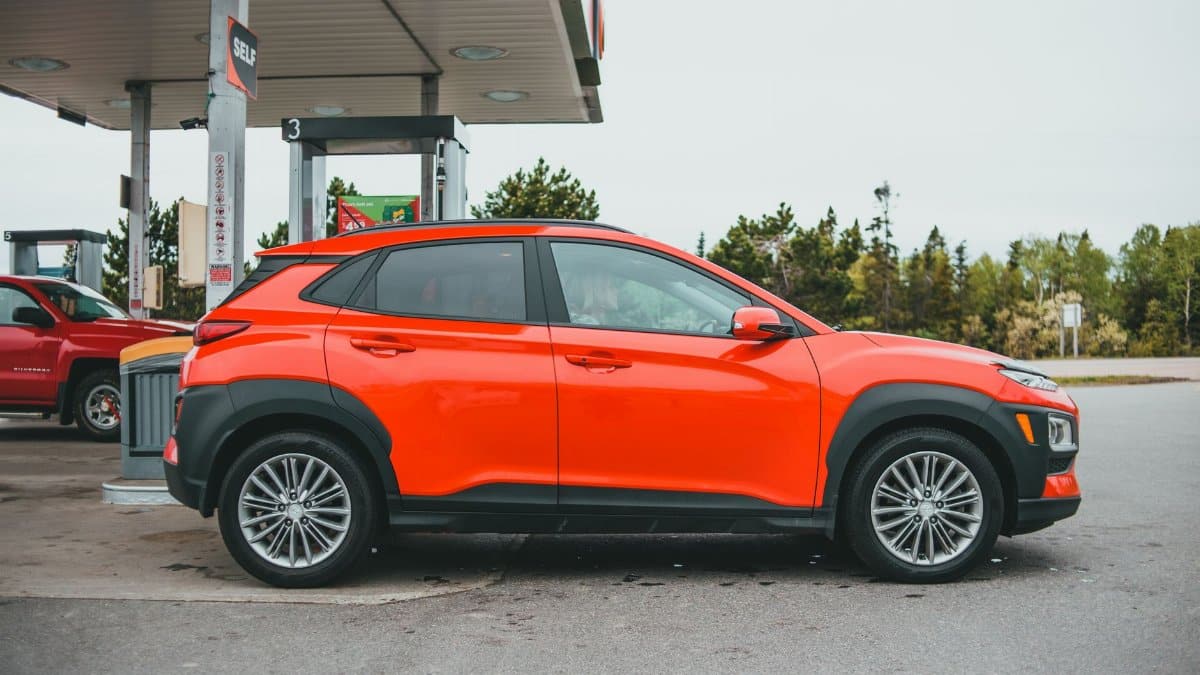Bright orange SUV parked at a gasoline station with a red truck in the background.