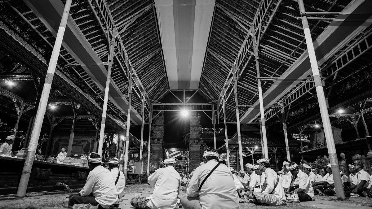 A gathering of men in traditional dress attending a night ceremony in a spacious open hall.