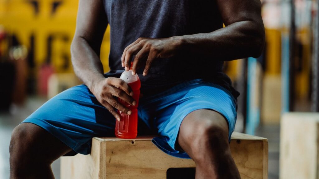 A man sitting on a wooden box, holding a red sports drink, taking a break from exercise.