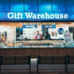 Night view of Gift Warehouse store with vibrant blue signage and two people inside.